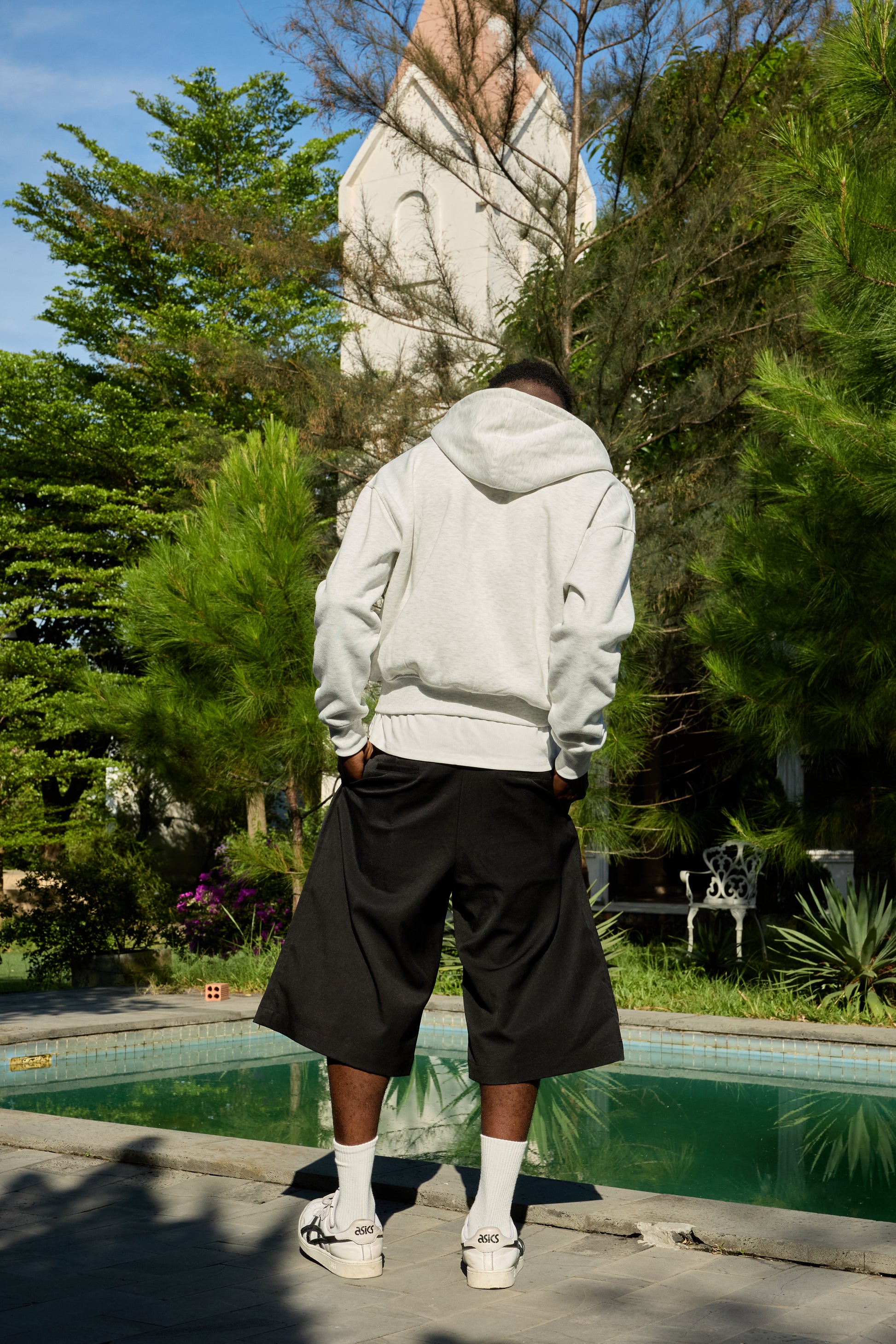 Person wearing a white hoodie and black shorts standing by a pool with trees and a house in the background.