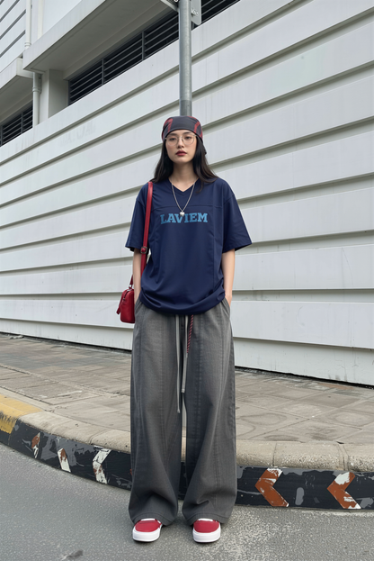 Person wearing a navy blue t-shirt with 'LAVIEM' text, gray wide-leg pants, and red shoes, standing against a white wall.