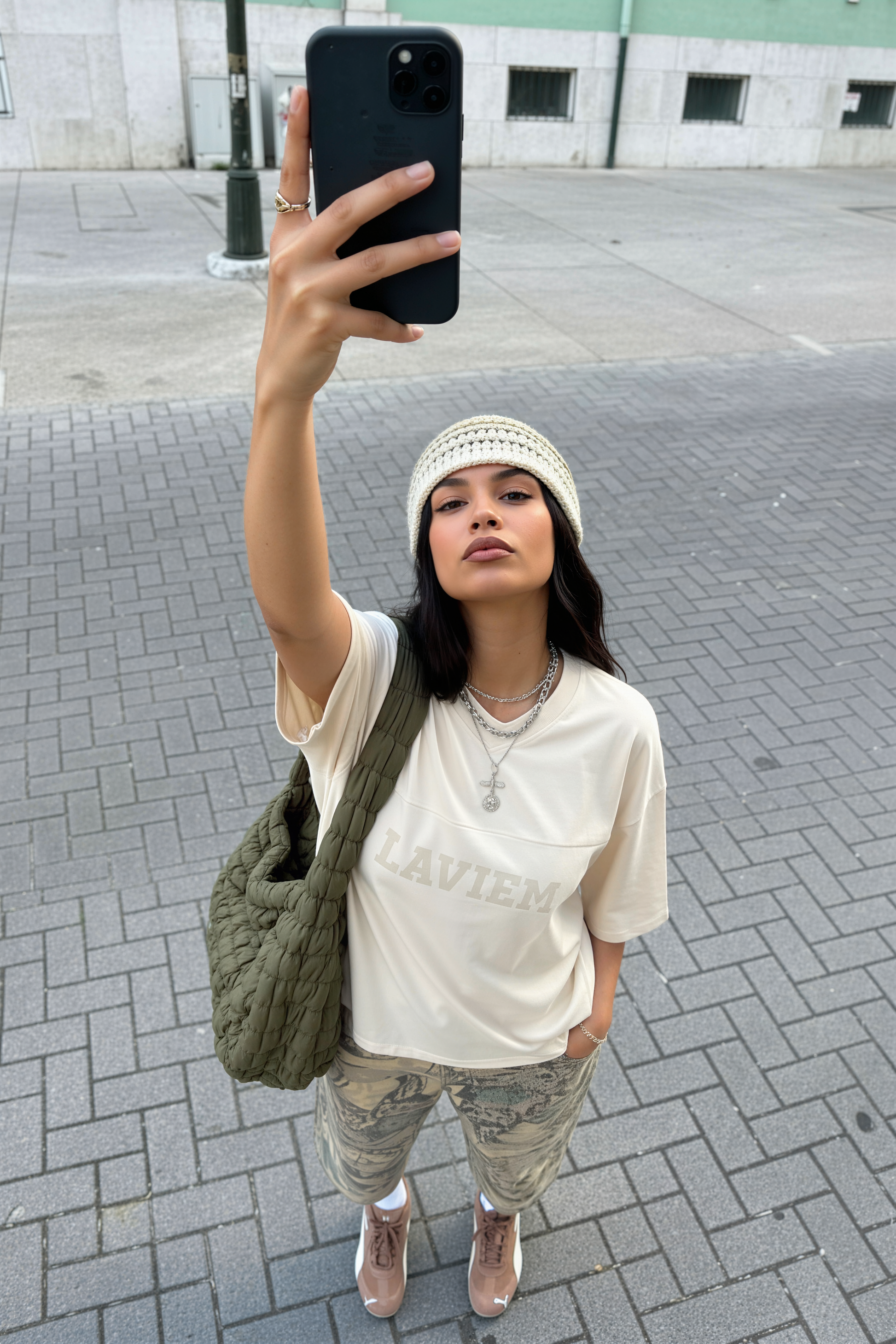 Woman taking a selfie on a street, wearing a white top, patterned pants, and a green bag.