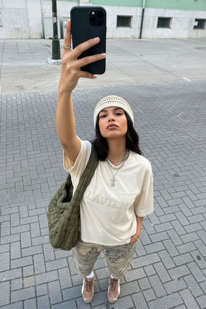 Woman taking a selfie on a street, wearing a white top, patterned pants, and a green bag.
