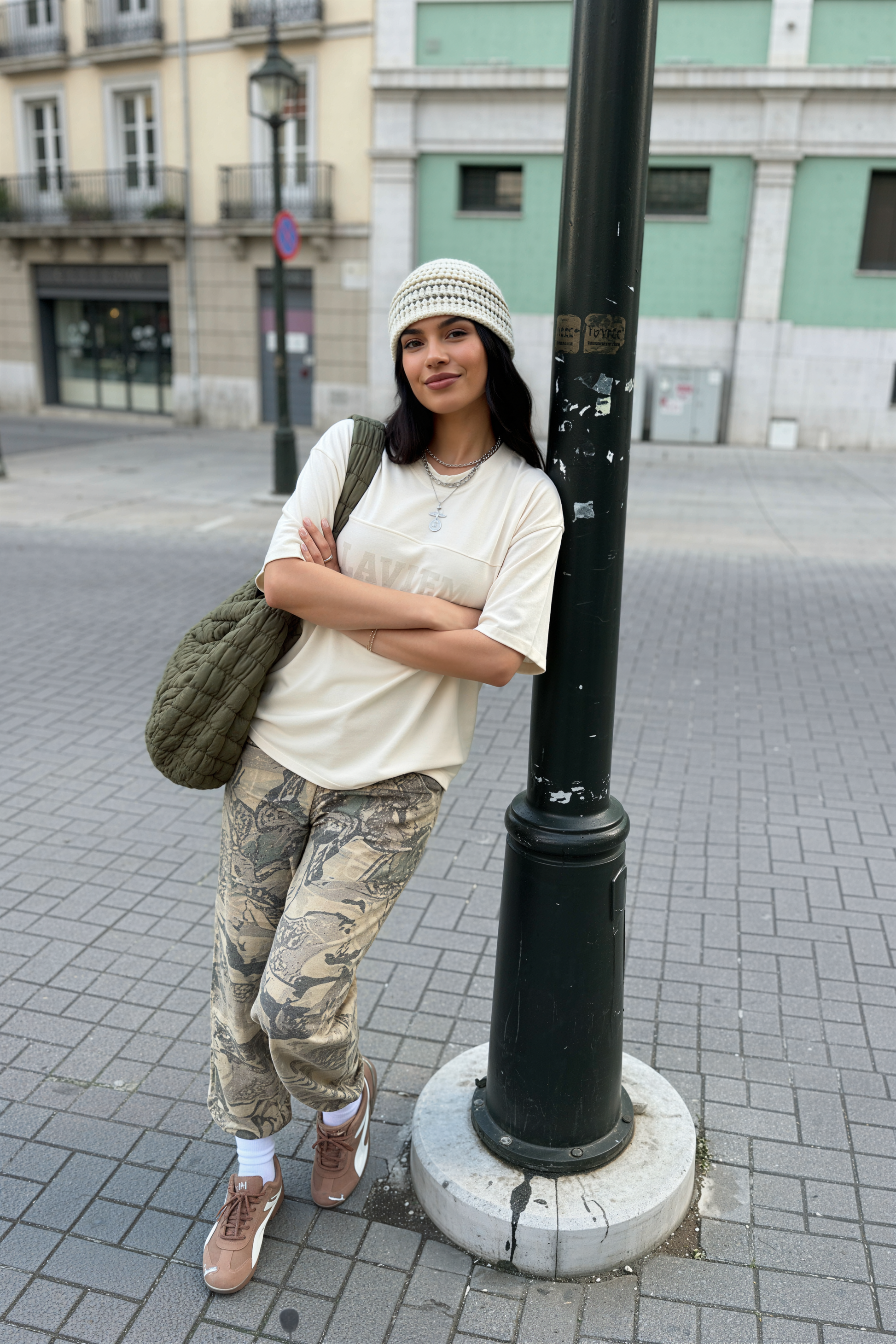 Woman standing on a street corner wearing a white shirt, camouflage pants, and a beanie.