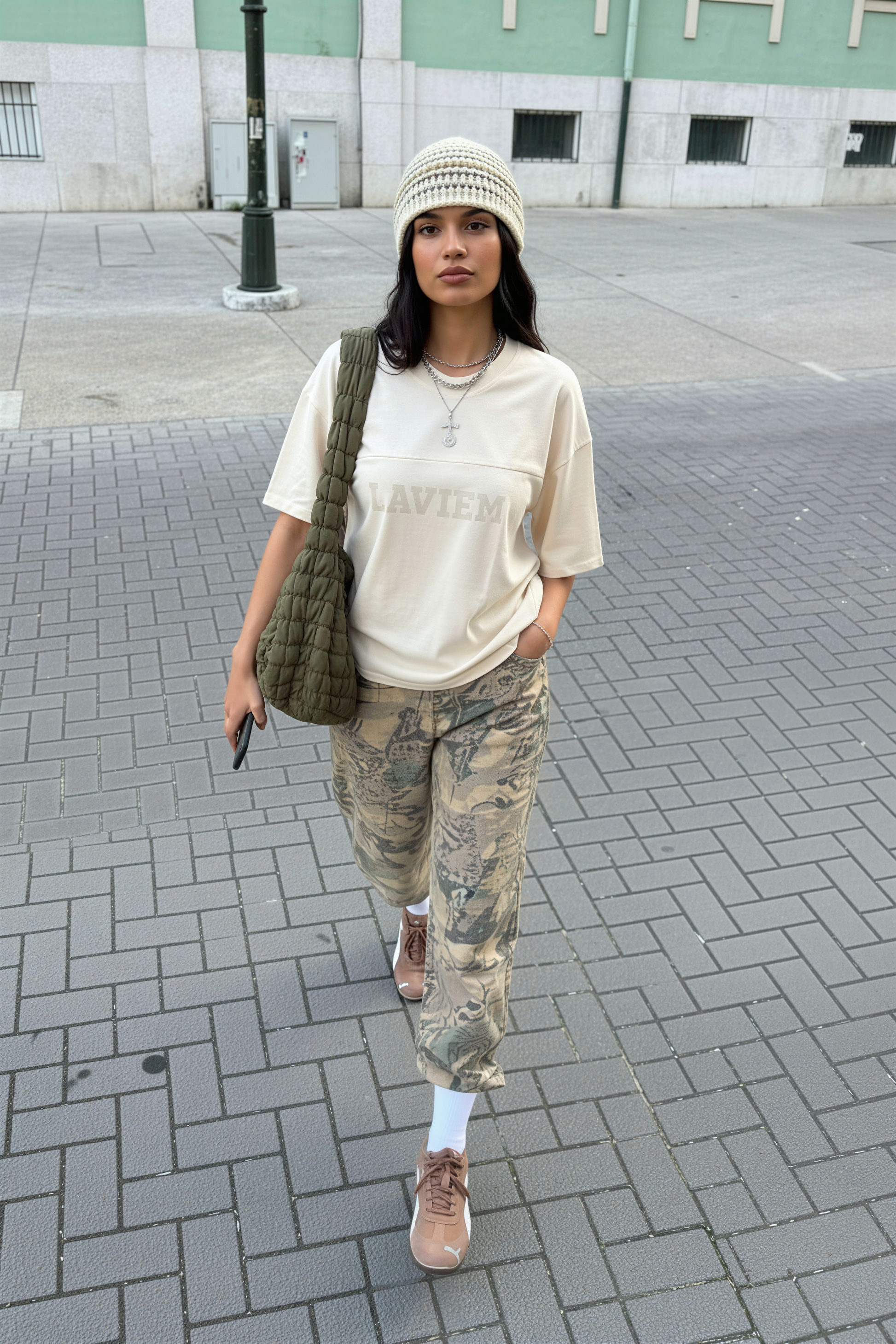 Person wearing a beige t-shirt, camouflage pants, and a green bag on a city street.