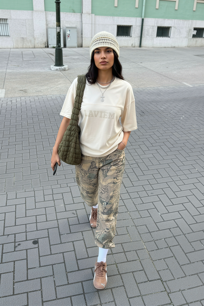 Person wearing a beige t-shirt, camouflage pants, and a green bag on a city street.